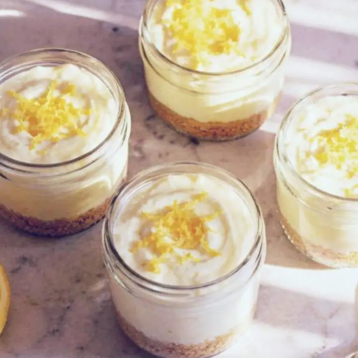 Four No-Bake Keto Lemon Cheesecake Cups topped with fresh lemon zest, served in small mason jars as a creamy low-carb dessert.